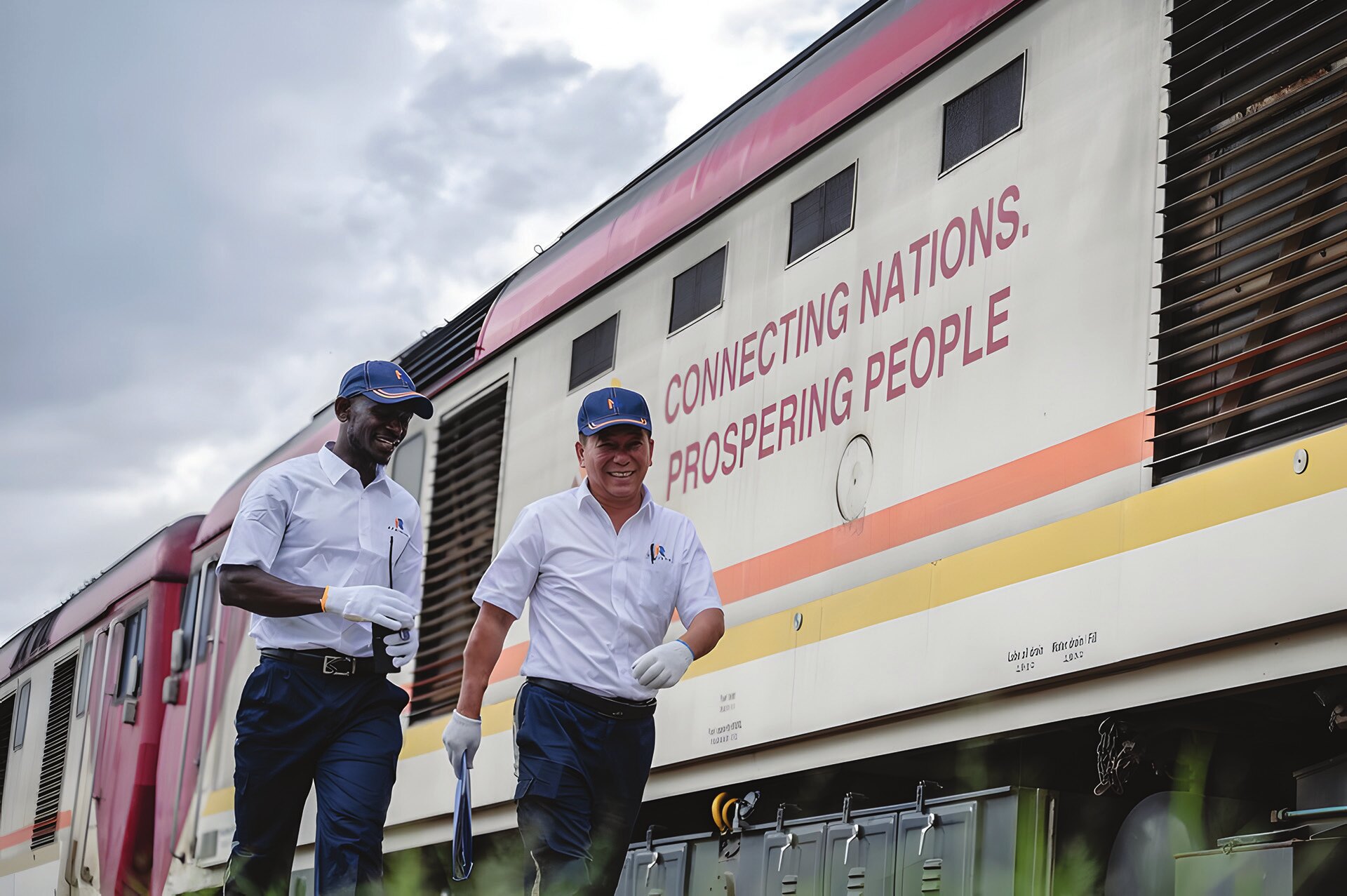Çinli eğitmen Jiang Liping (sağda) ve stajyer Horace Owiti, 23 Mayıs 2023'te Nairobi, Kenya'da Mombasa-Nairobi Demiryolu üzerinde “Ulusları birbirine bağlıyor, halkı zenginleştiriyor.” yazılı bir tren vagonunun yanından geçerken (Fotoğraf: Wang Guansen, Xinhua, 2023).